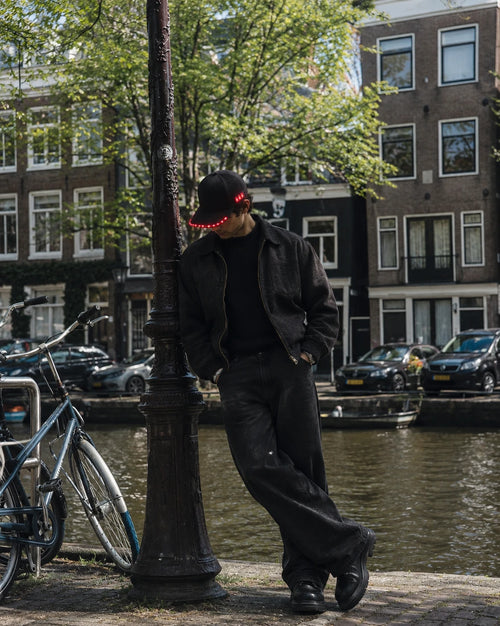 Man leaning on a lamppost by an Amsterdam canal, wearing the Wellora cap with its red LEDs lit